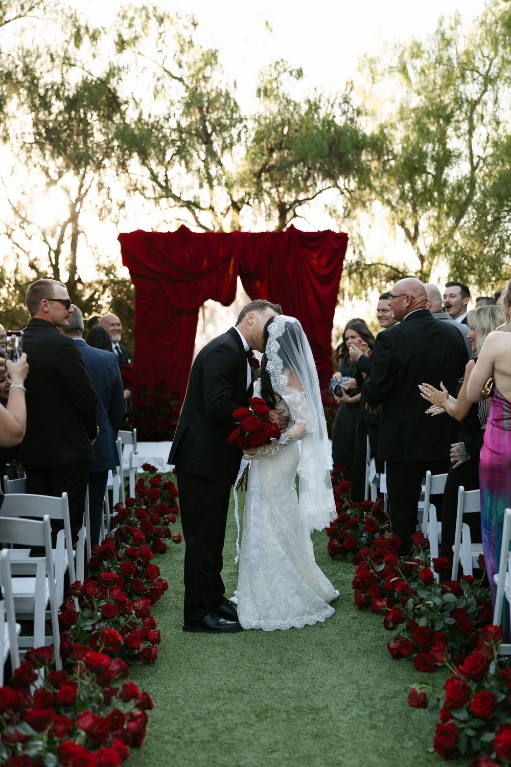 Bride and groom share first kiss at outdoor wedding ceremony with red rose aisle and dramatic velvet backdrop.