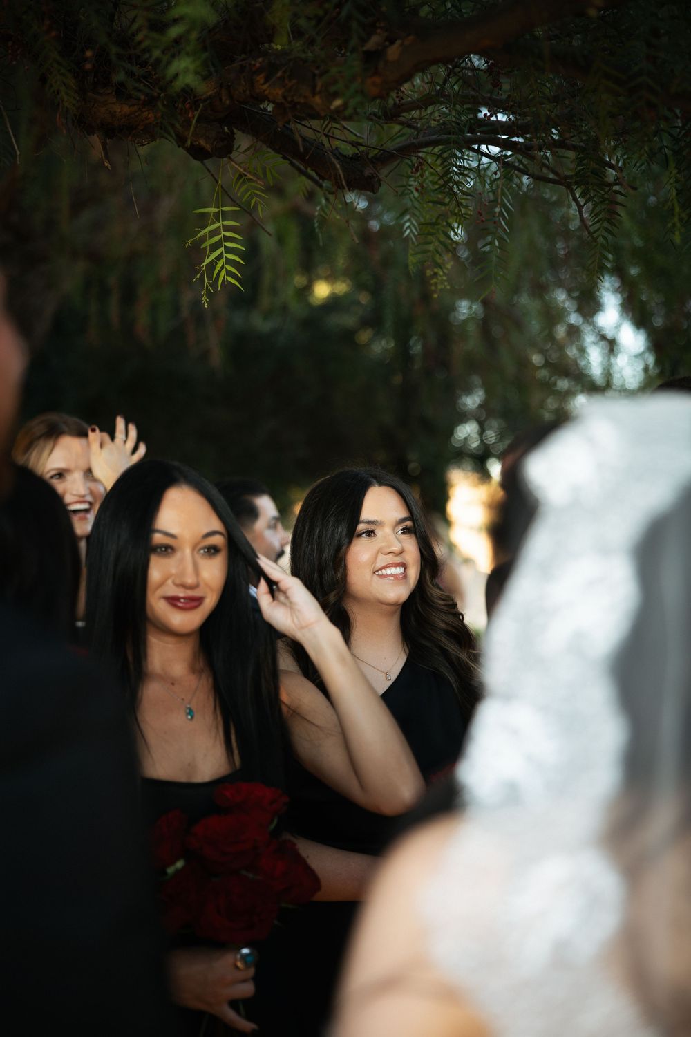 Bridesmaids in black dresses holding red roses smile warmly during an outdoor wedding ceremony surrounded by lush greenery.