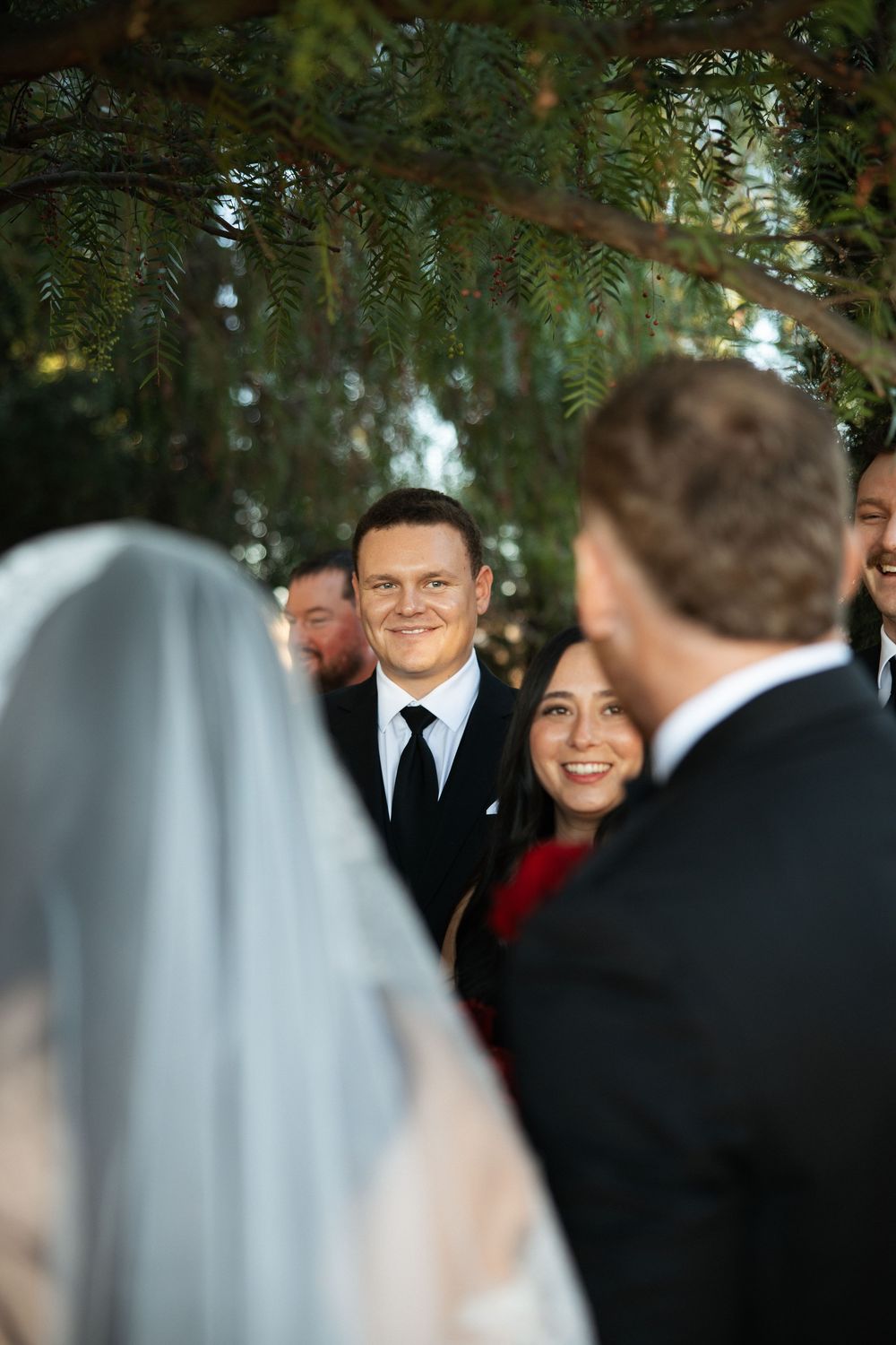 Groom smiling joyfully at bride during outdoor wedding ceremony with guests watching under green tree canopy.