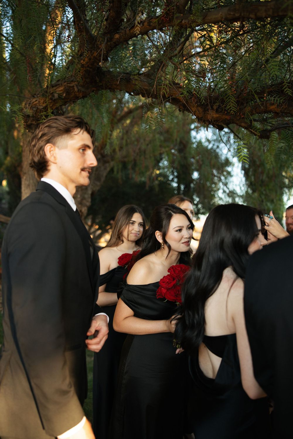 Elegant guests in black formal attire mingle outdoors under pine trees at an evening event.