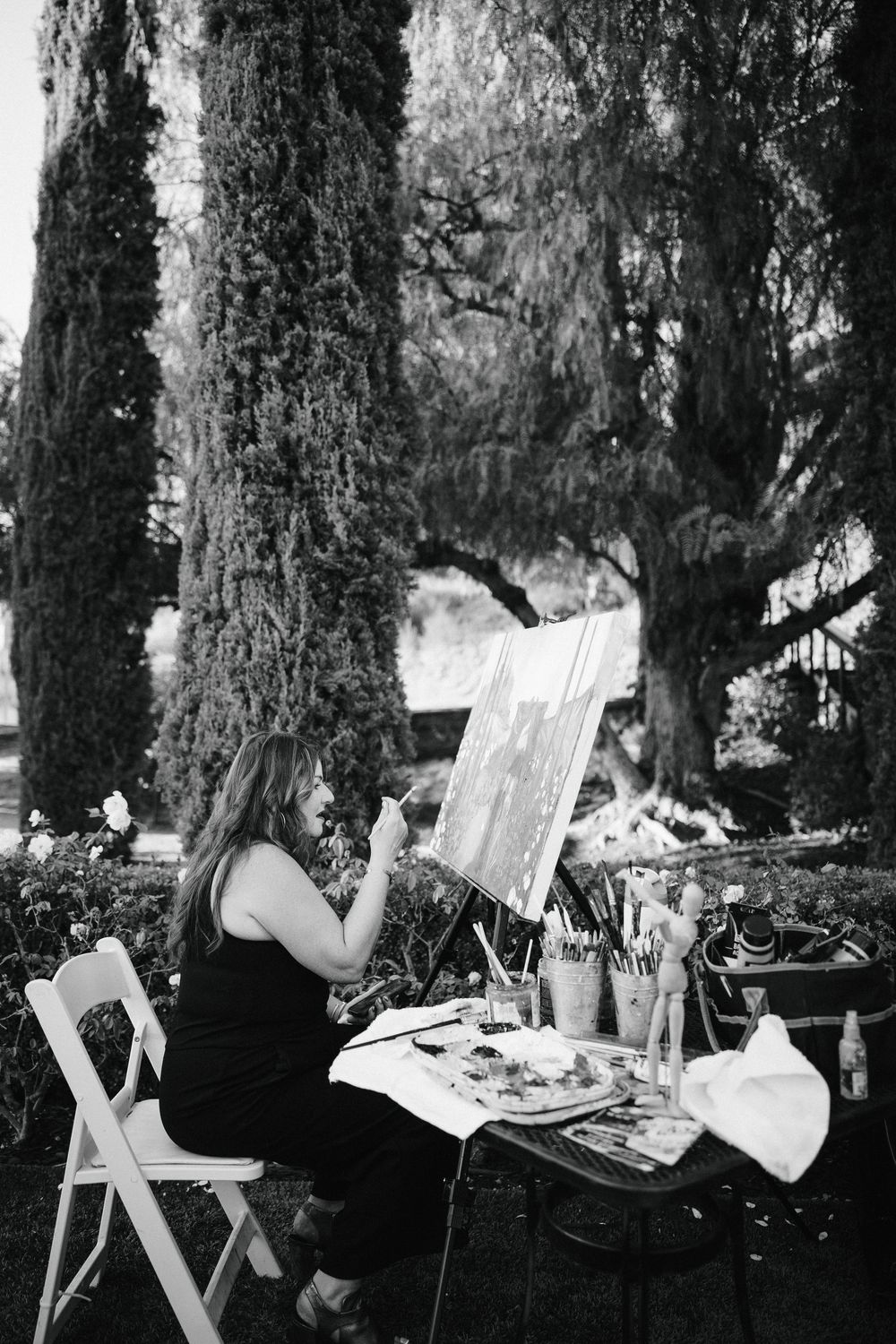 Woman in black dress painting on easel outdoors surrounded by cypress trees with art supplies on table nearby.