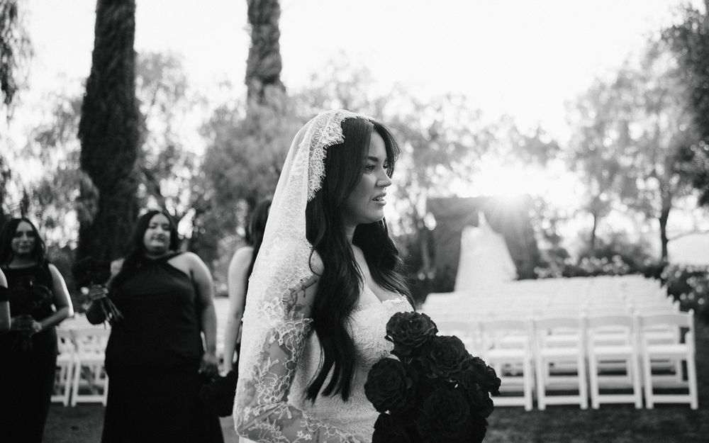 Bride in lace wedding dress and veil holding red roses bouquet at outdoor ceremony with golden sunset backdrop.