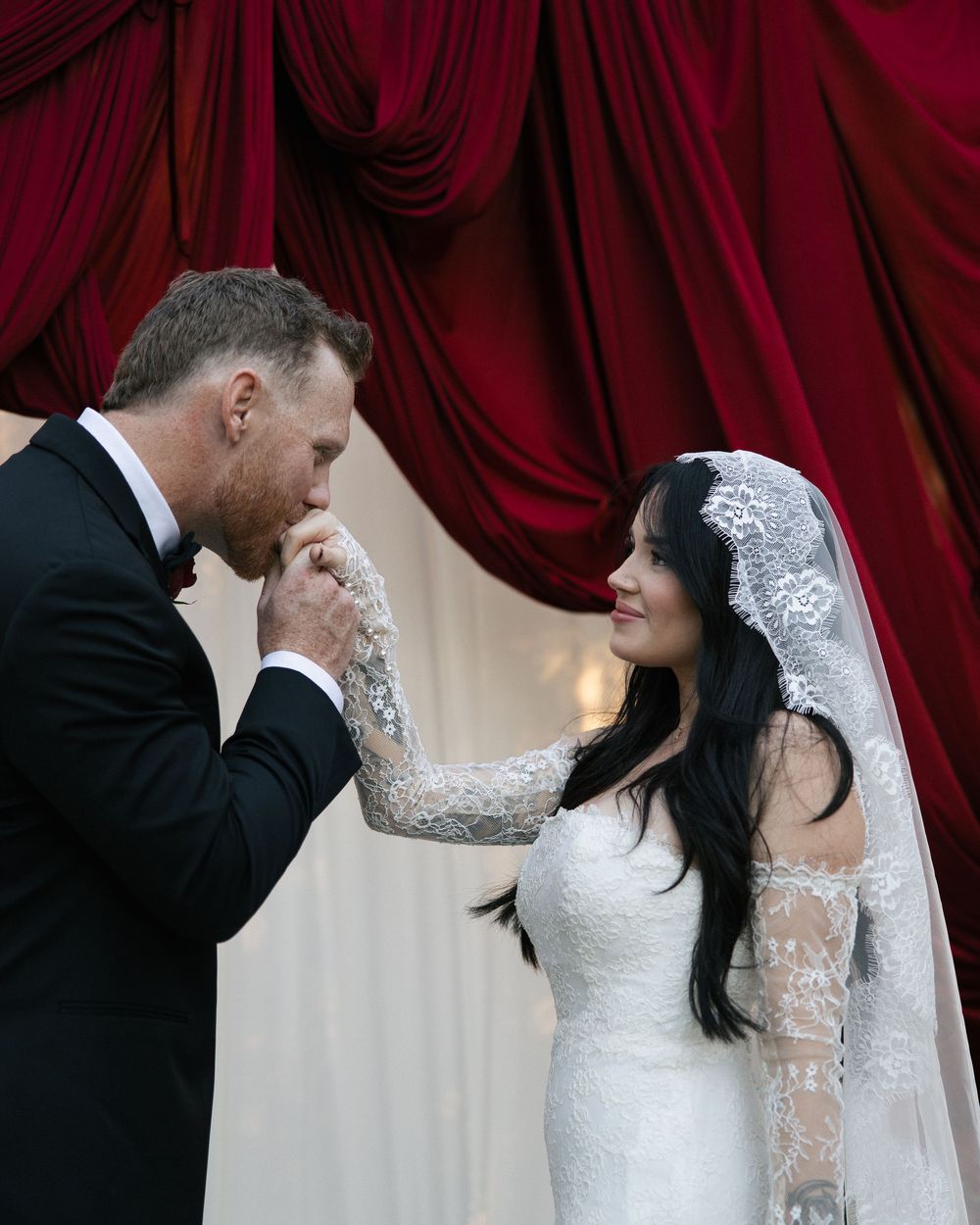 Groom in black tuxedo kissing bride's hand at altar with red velvet drapes, bride in lace gown and veil smiling.