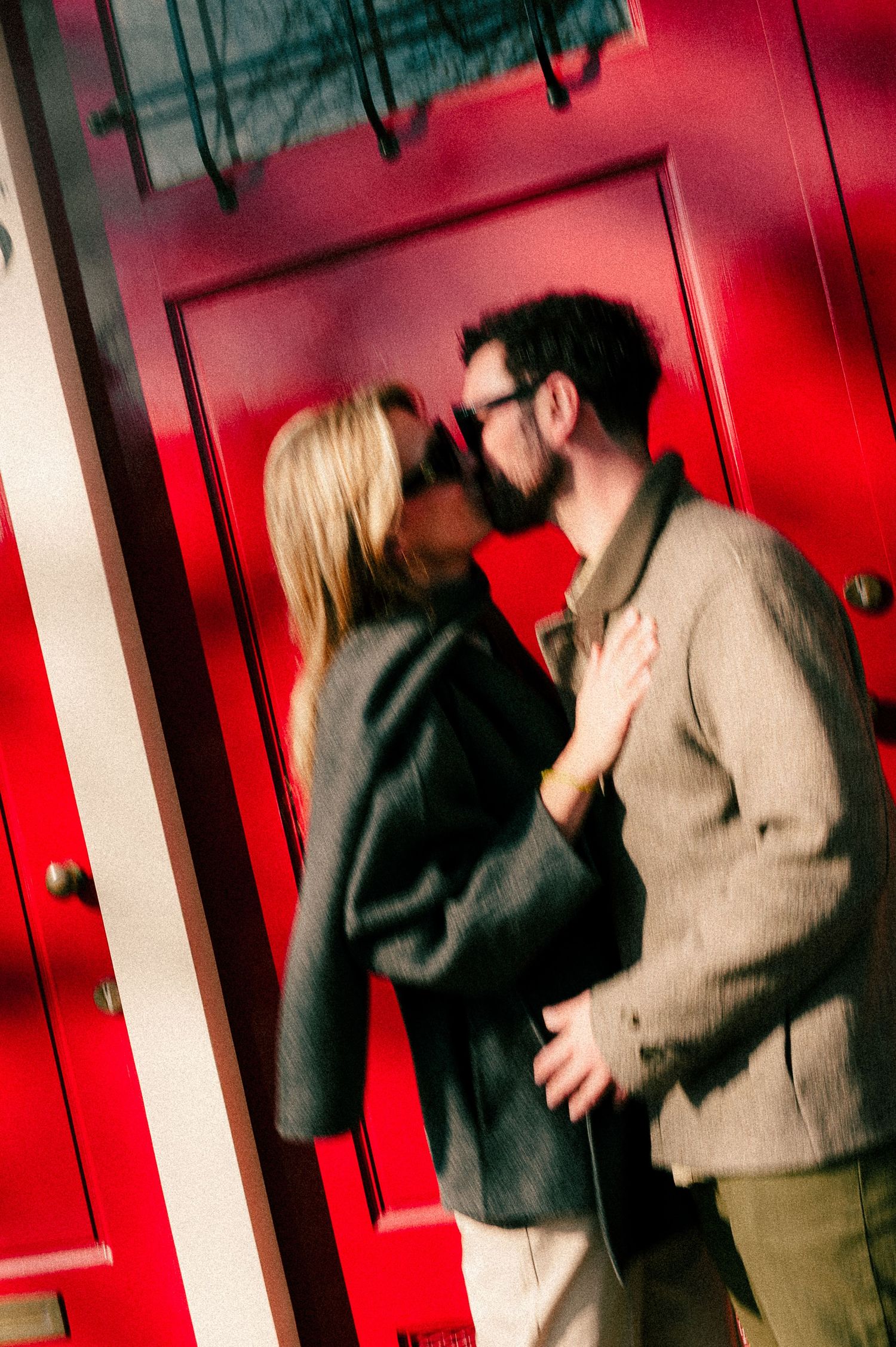 A couple sharing a romantic kiss in front of vibrant red doors, woman in black blazer and man in beige jacket wearing glasses.