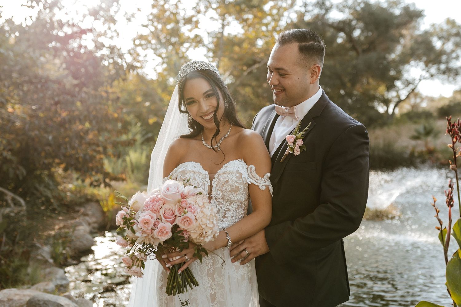 Bride and groom pose together outdoors near a pond, she holds a pink bouquet in a lace gown with veil.