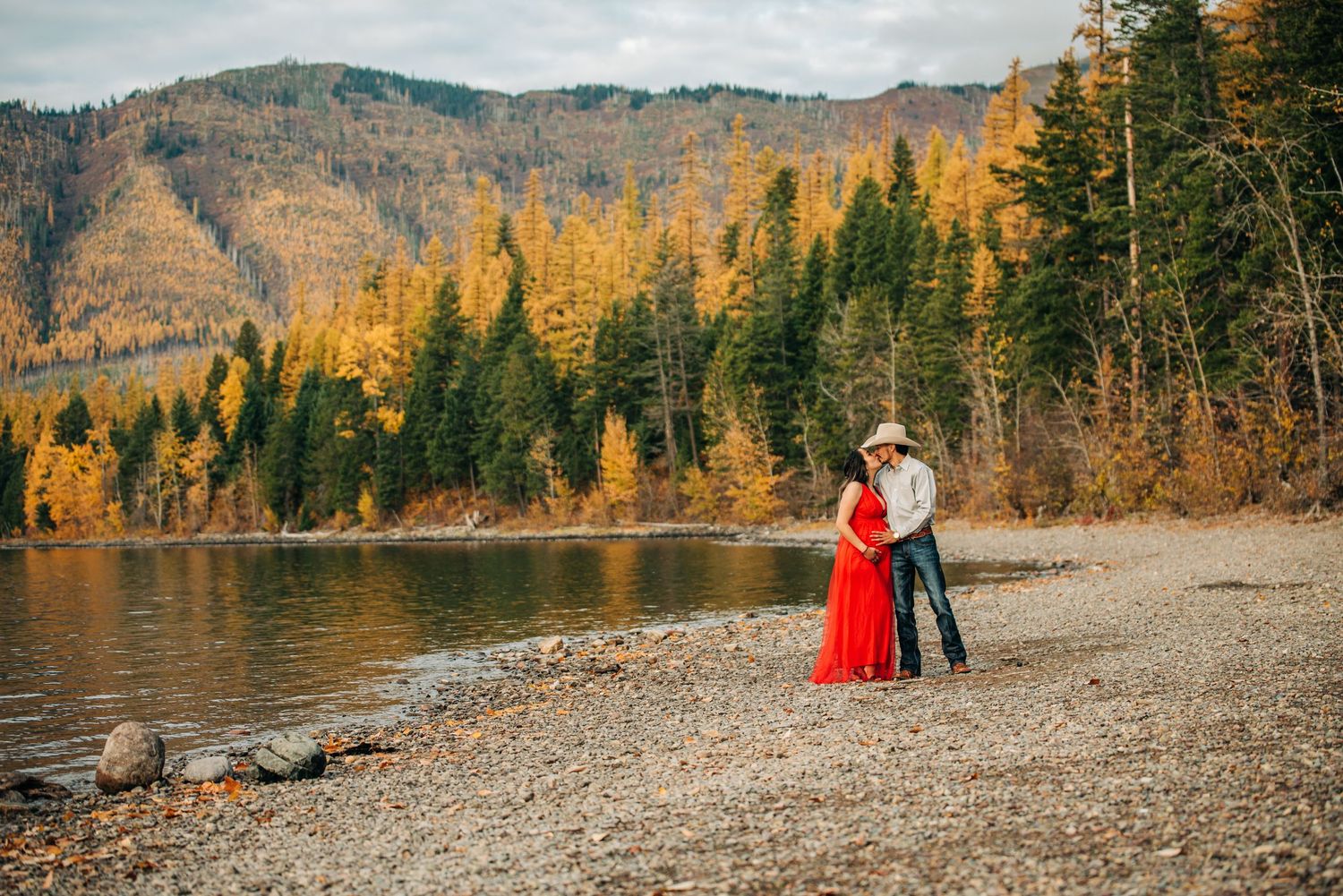 Couple embracing on rocky lakeshore with golden autumn trees and mountain backdrop in fall season.