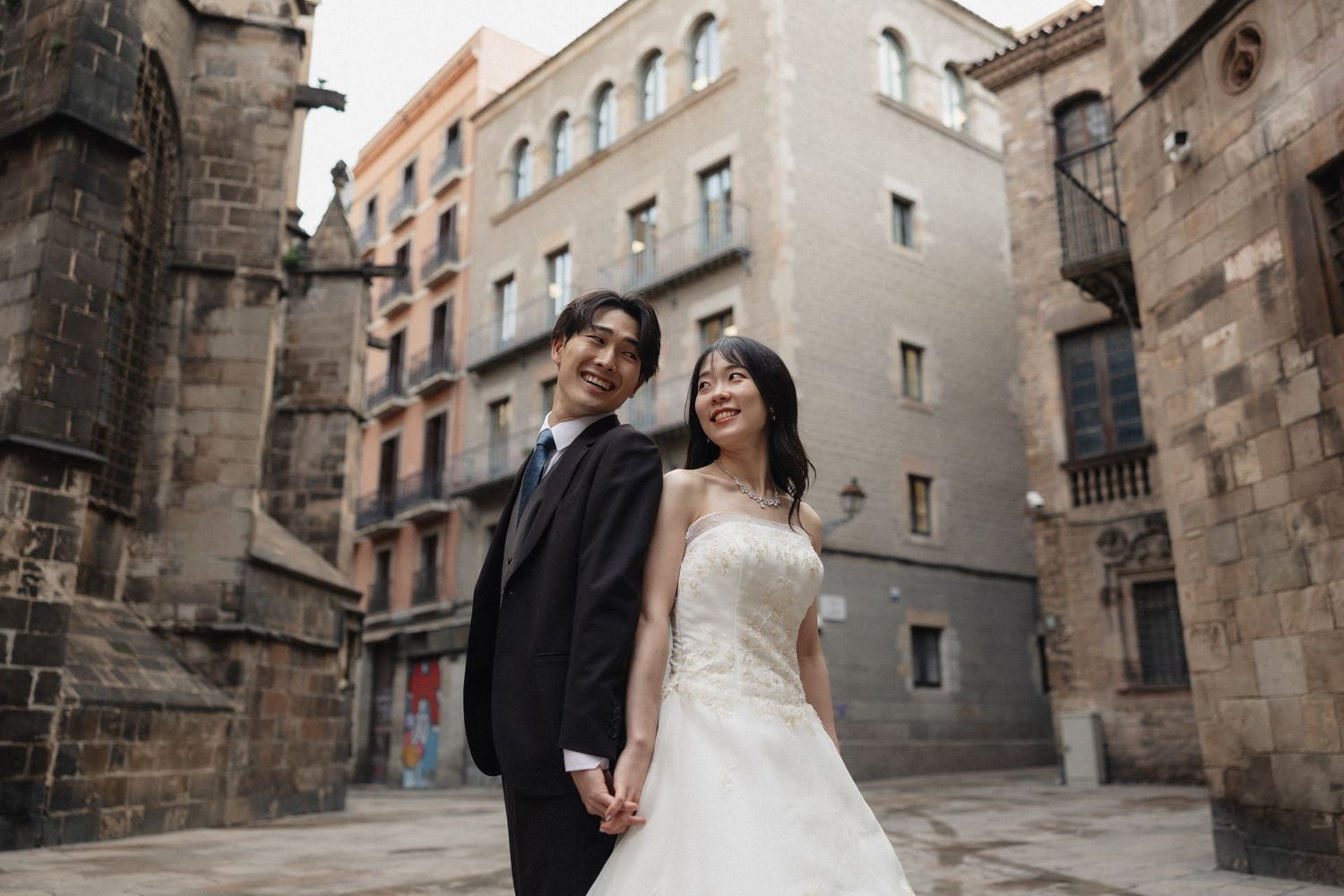 A couple in wedding attire stands together in front of historic stone buildings in a European city street.
