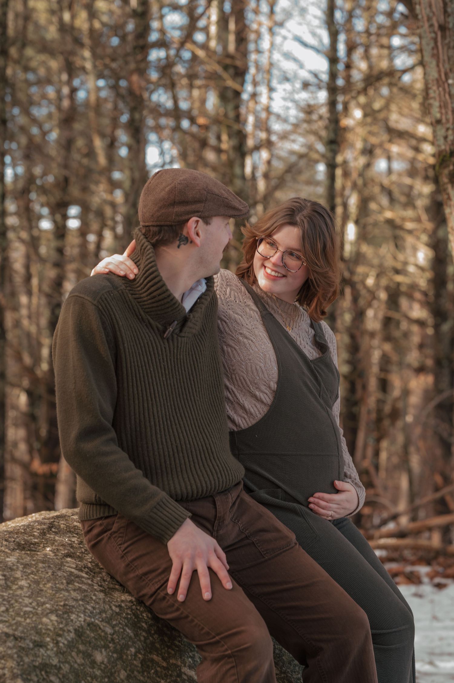Couple sitting on a rock in a winter forest, woman visibly pregnant, both smiling warmly in earth-toned clothing.