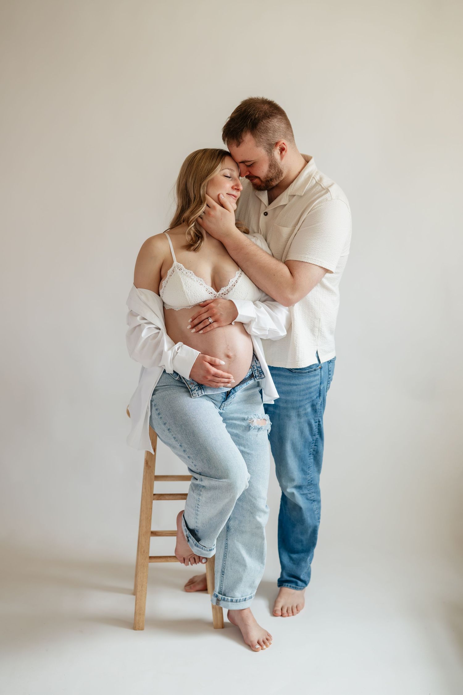 Intimate maternity photography session showing tender moments between couple in casual white and denim against neutral backdrop.