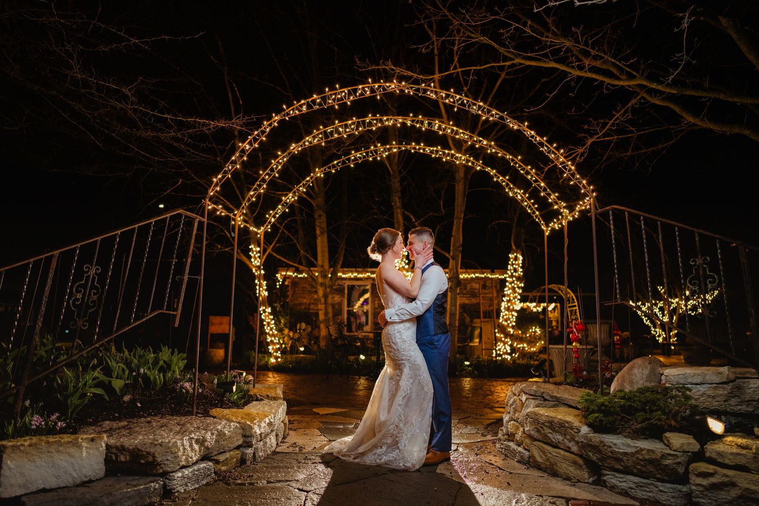 Couple sharing romantic kiss under glowing arch of fairy lights at night during outdoor wedding celebration.
