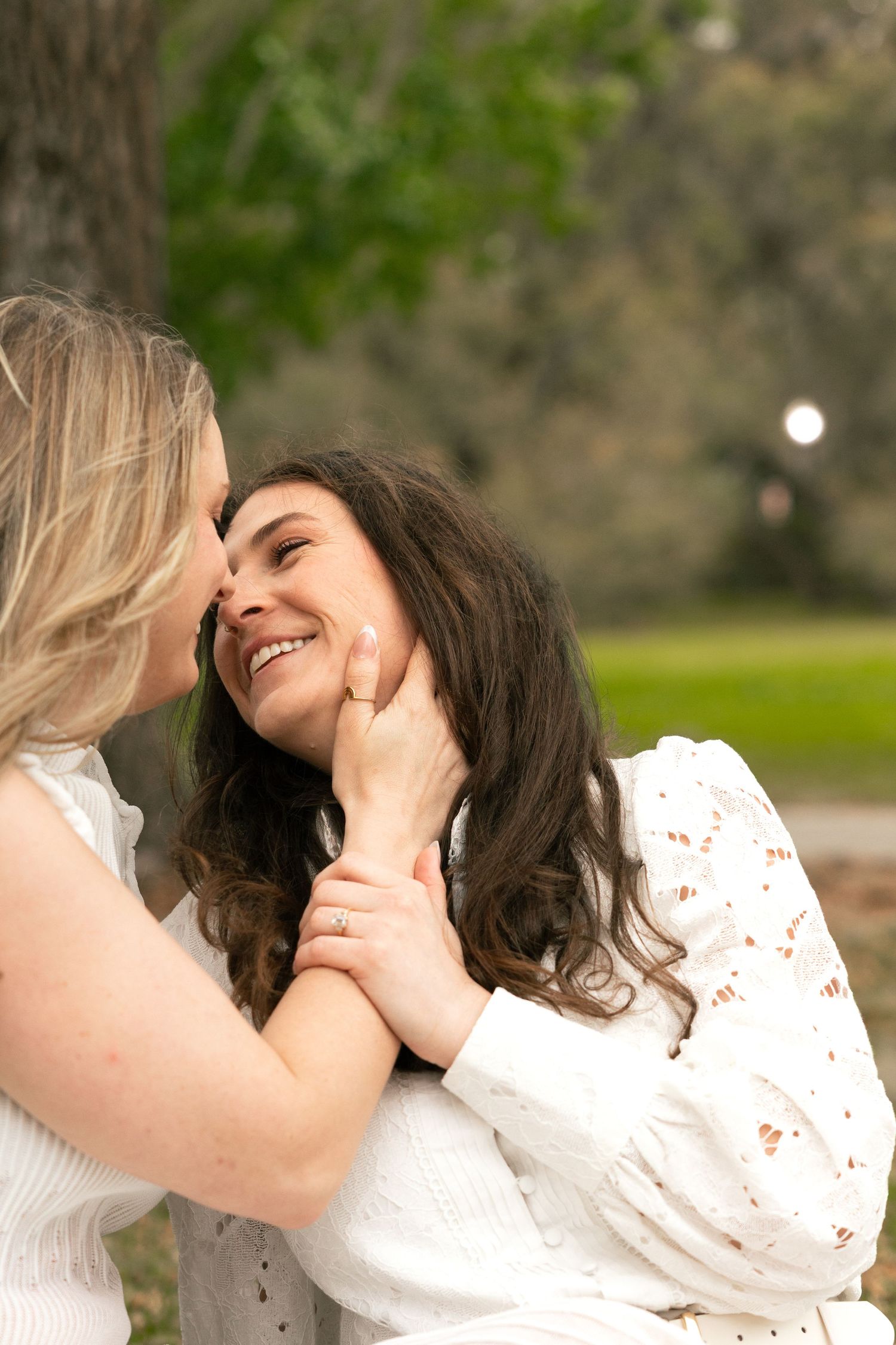 Two women in white dresses share a tender moment outdoors, one smiling as the other gently holds her face.