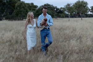 Father holding new born and mother holding onto father while walking through a paddock during a professional newborn photo shoot.