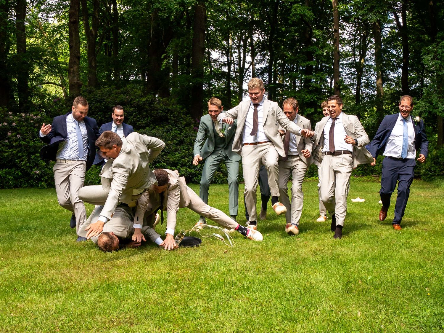 Bruidsjonkers in beige pakken dollen op elkaar op een groen gazon omringd door bomen.