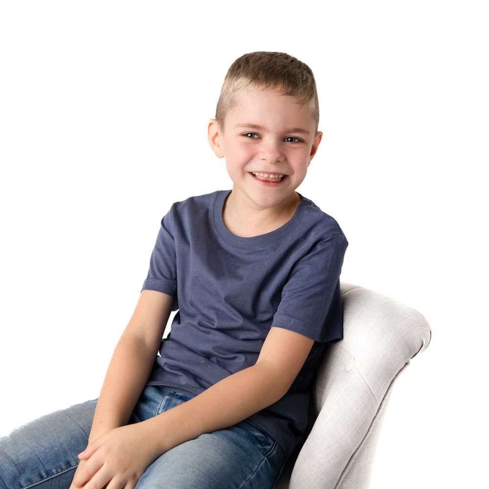 Smiling young boy in navy t-shirt and jeans sitting in a white chair against a white background.