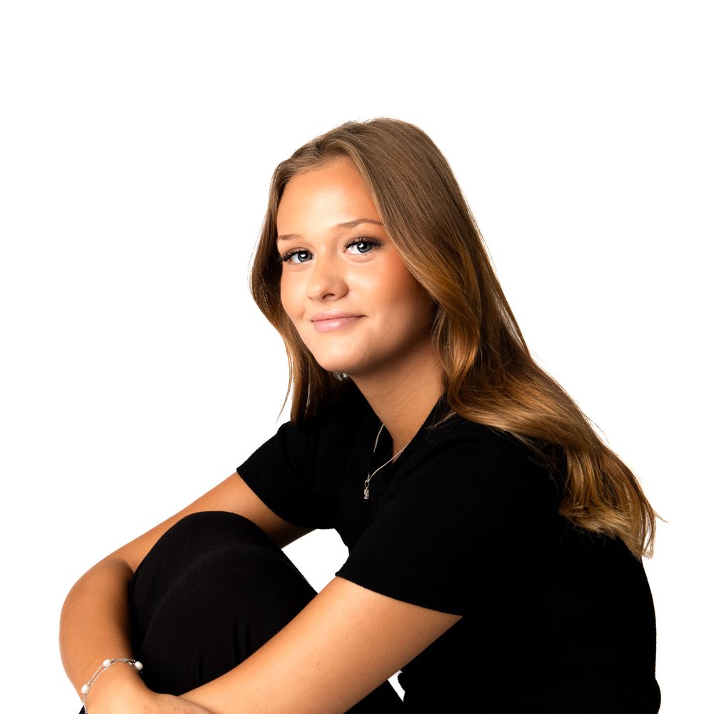Young teenage girl with long brown hair wearing a black top, smiling confidently against a white background.