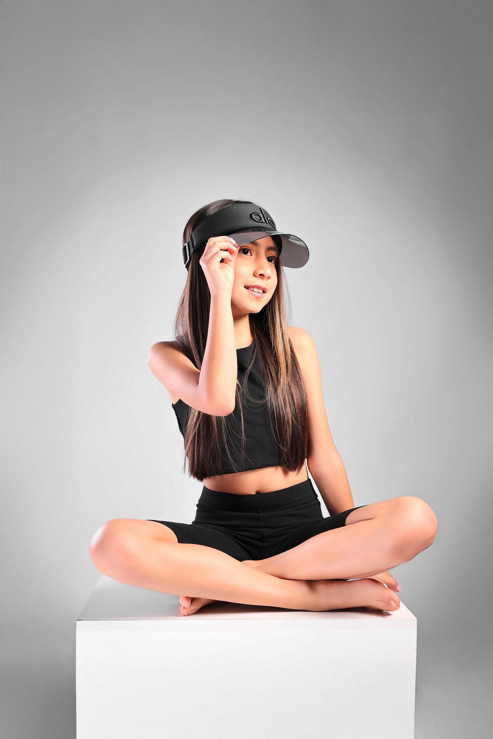 Young woman in black crop top and shorts sitting cross-legged on white cube, adjusting black cap, studio shot.