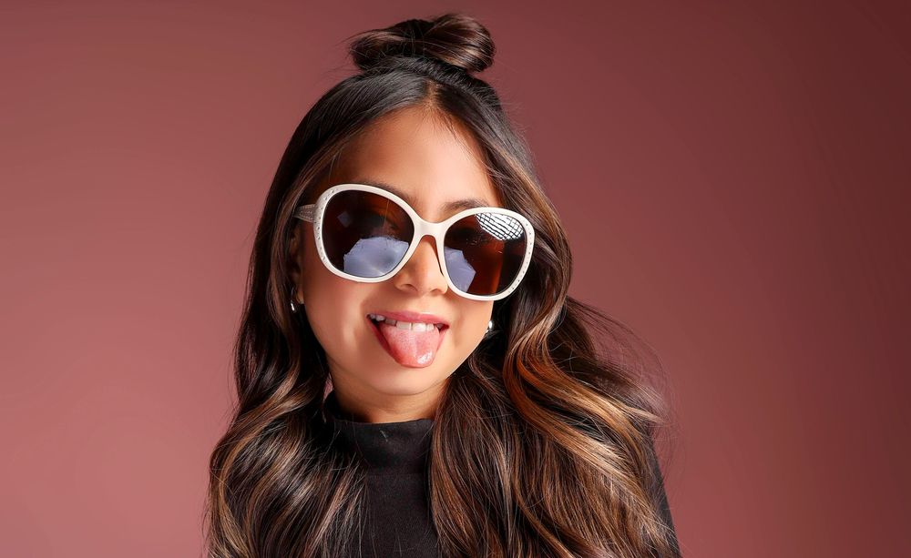 Young woman wearing oversized white sunglasses, sticking tongue out, with long dark hair, against mauve background.