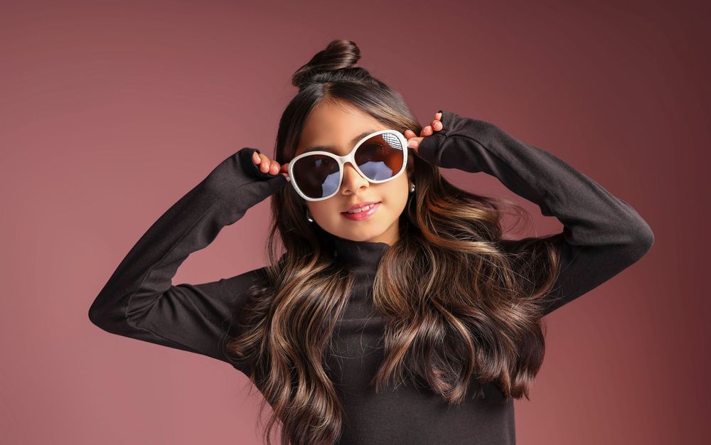 Young girl posing with oversized round sunglasses, wearing a black top, with long wavy hair against a pink background.