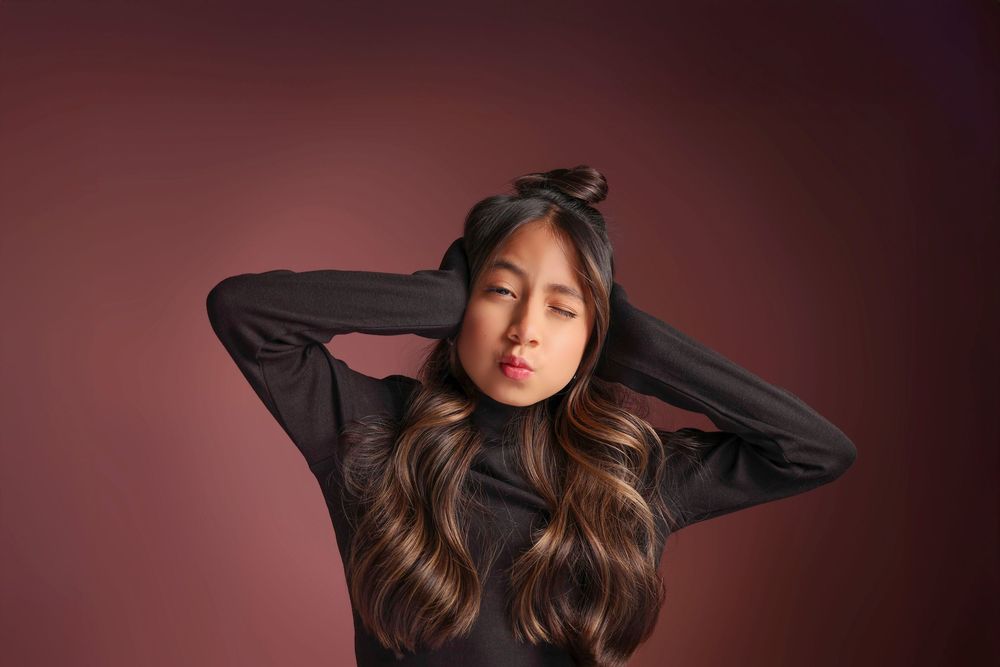 Young Asian woman with long highlighted hair poses in black turtleneck against dark red background.