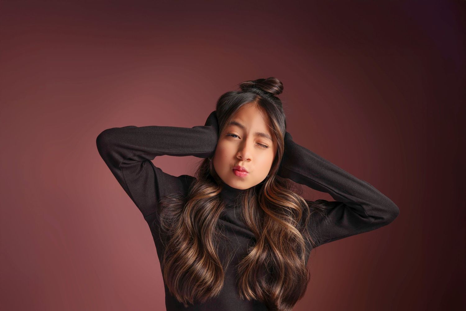 Young Asian woman with long highlighted hair poses in black turtleneck against dark red background.