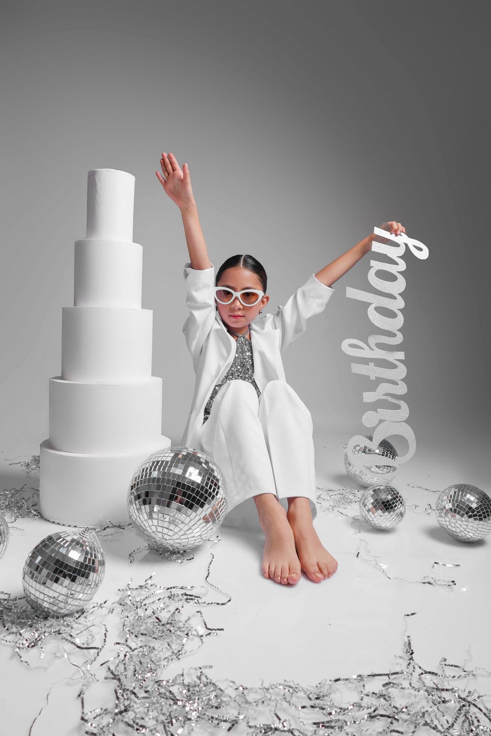 Young girl in white outfit with sunglasses poses by tiered cake, disco balls, and Birthday sign on gray background.