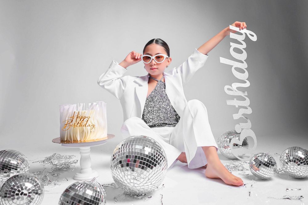 Woman in white suit with disco balls, birthday cake, and Birthday sign on white background.