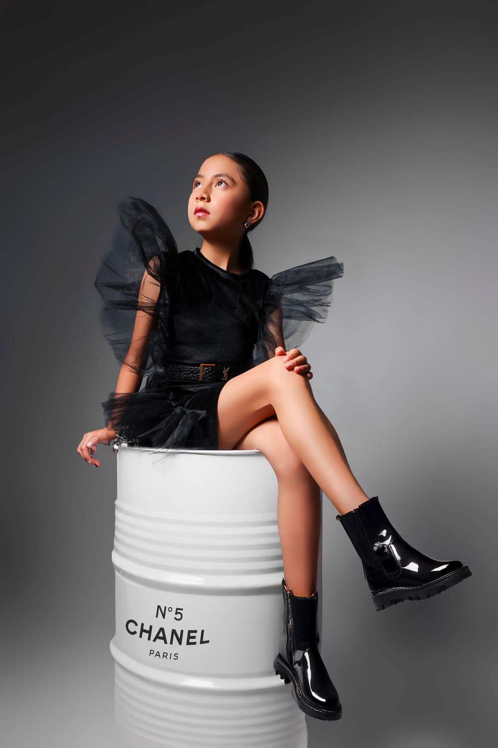 Young girl in black tulle dress with wings sits on a Chanel No.5 barrel, wearing patent boots.