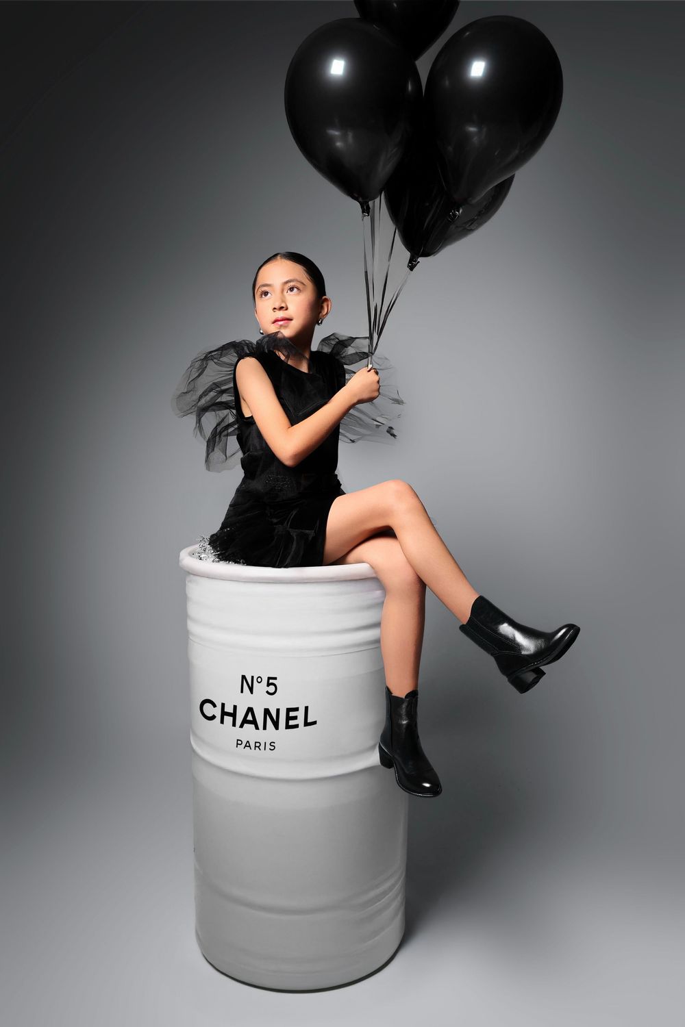 Young girl in black dress sitting on Chanel No.5 barrel holding black balloons in a fashion editorial shoot.