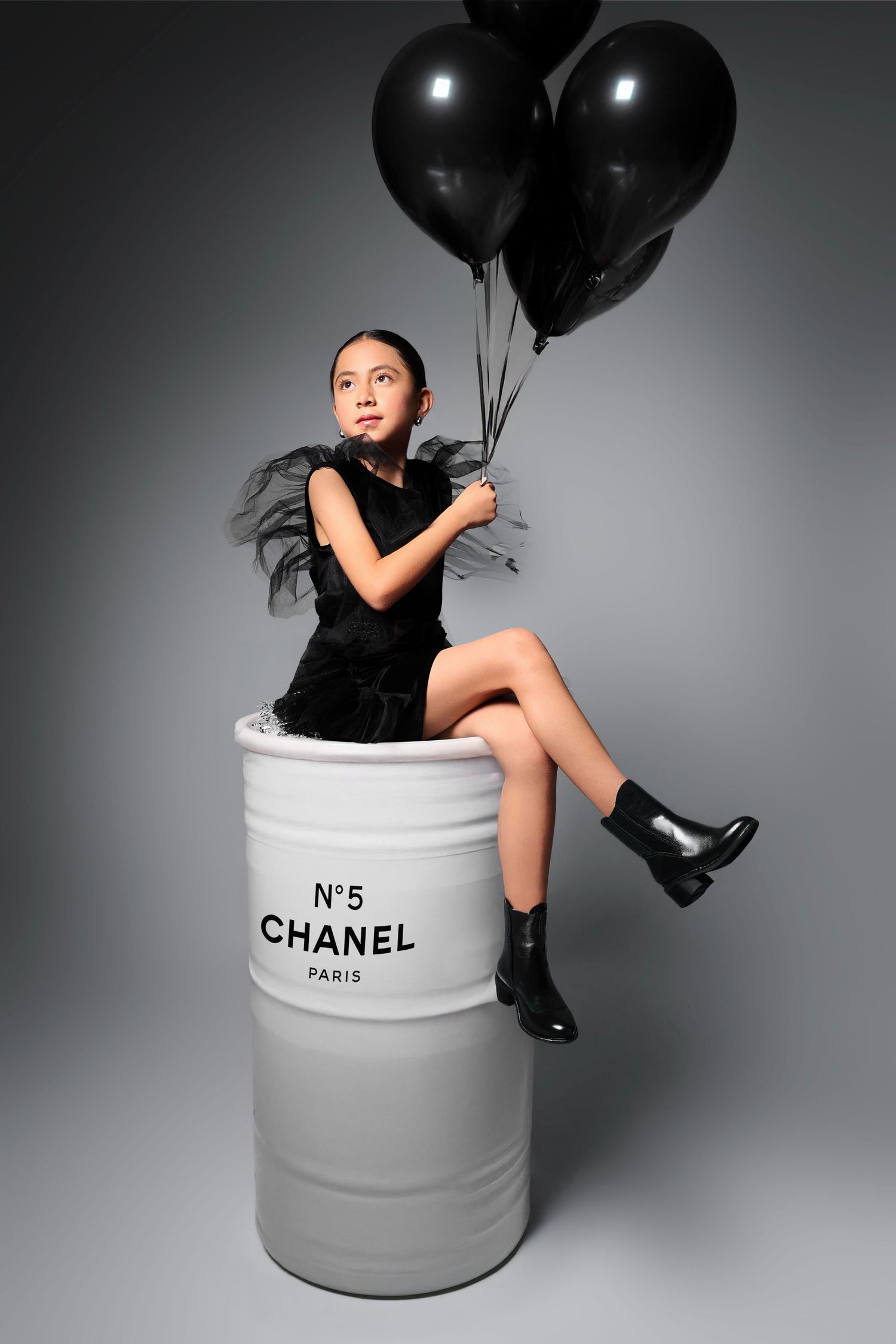 Young girl in black dress sitting on Chanel No.5 barrel holding black balloons in a fashion editorial shoot.