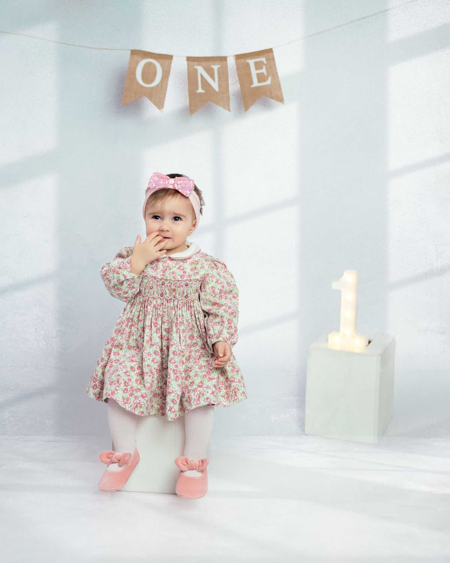 Baby girl in floral dress celebrating first birthday with ONE banner and number 1 block in soft studio.