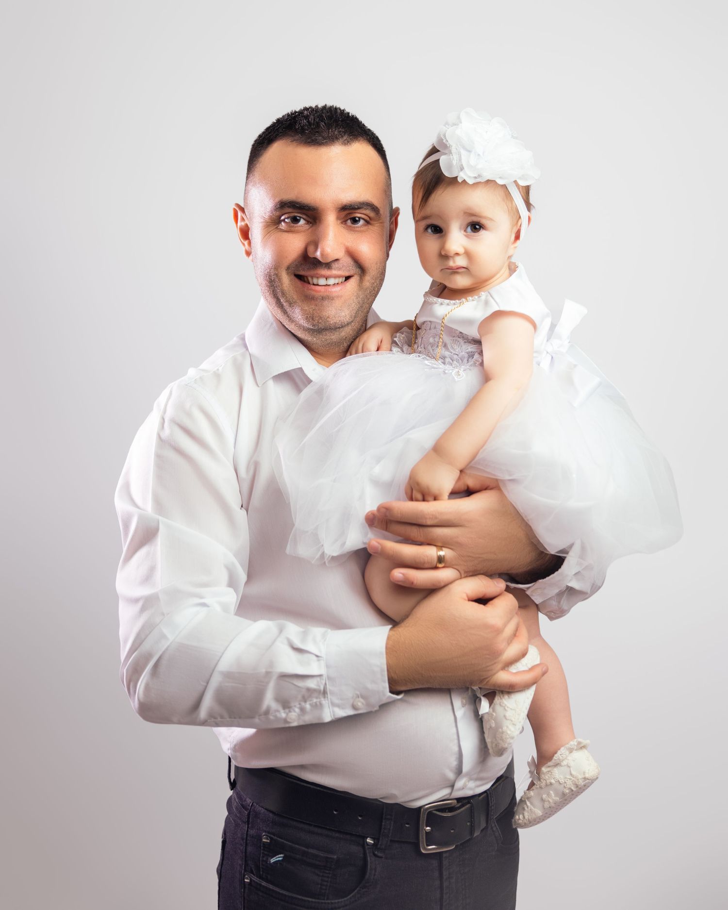 Smiling father holding baby daughter dressed in white lace outfit with bow headband in studio portrait.
