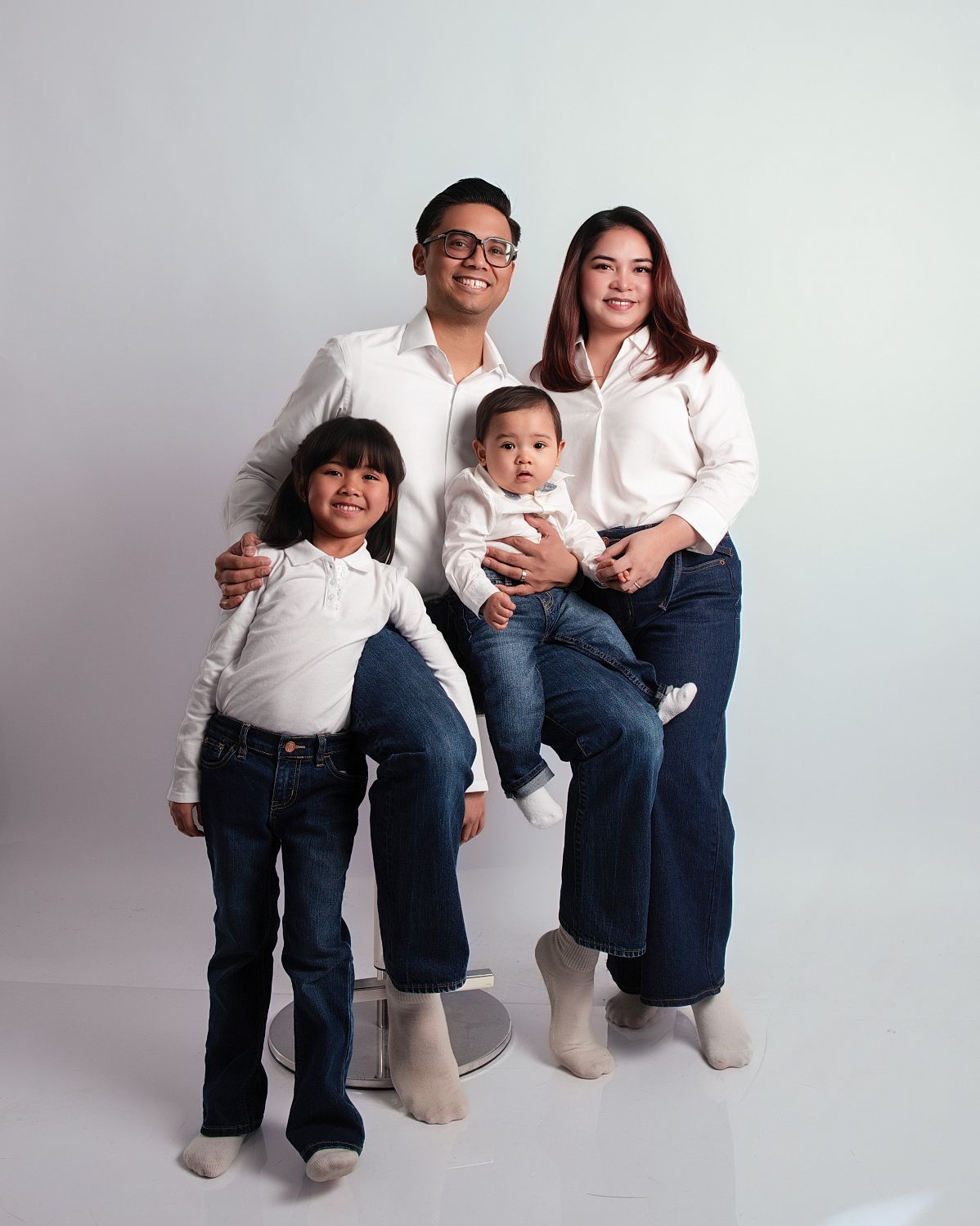Family of four in matching white shirts and jeans posing together in a bright studio portrait session.