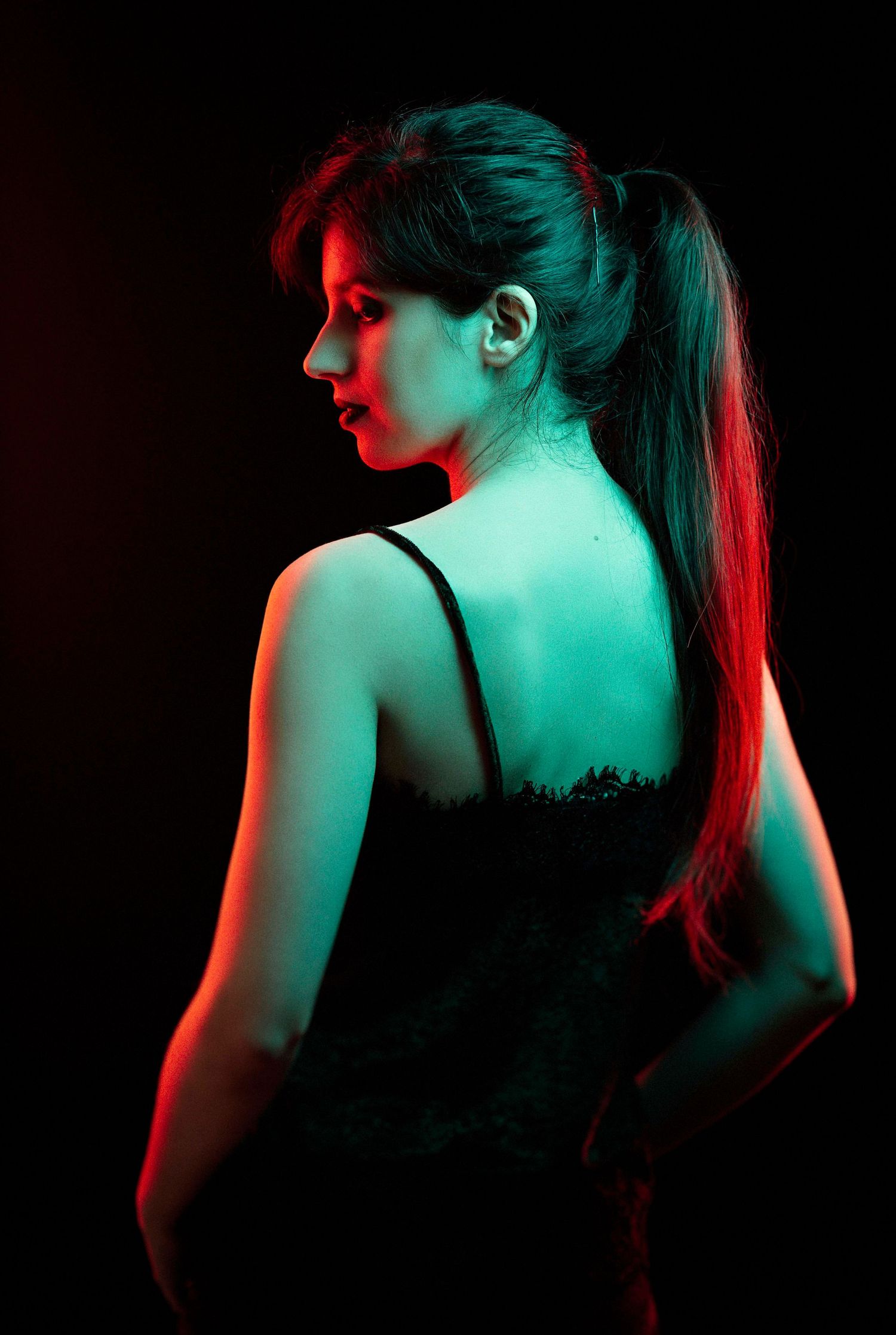 Woman in black lace top with ponytail posed against dark background with dramatic red and green lighting.