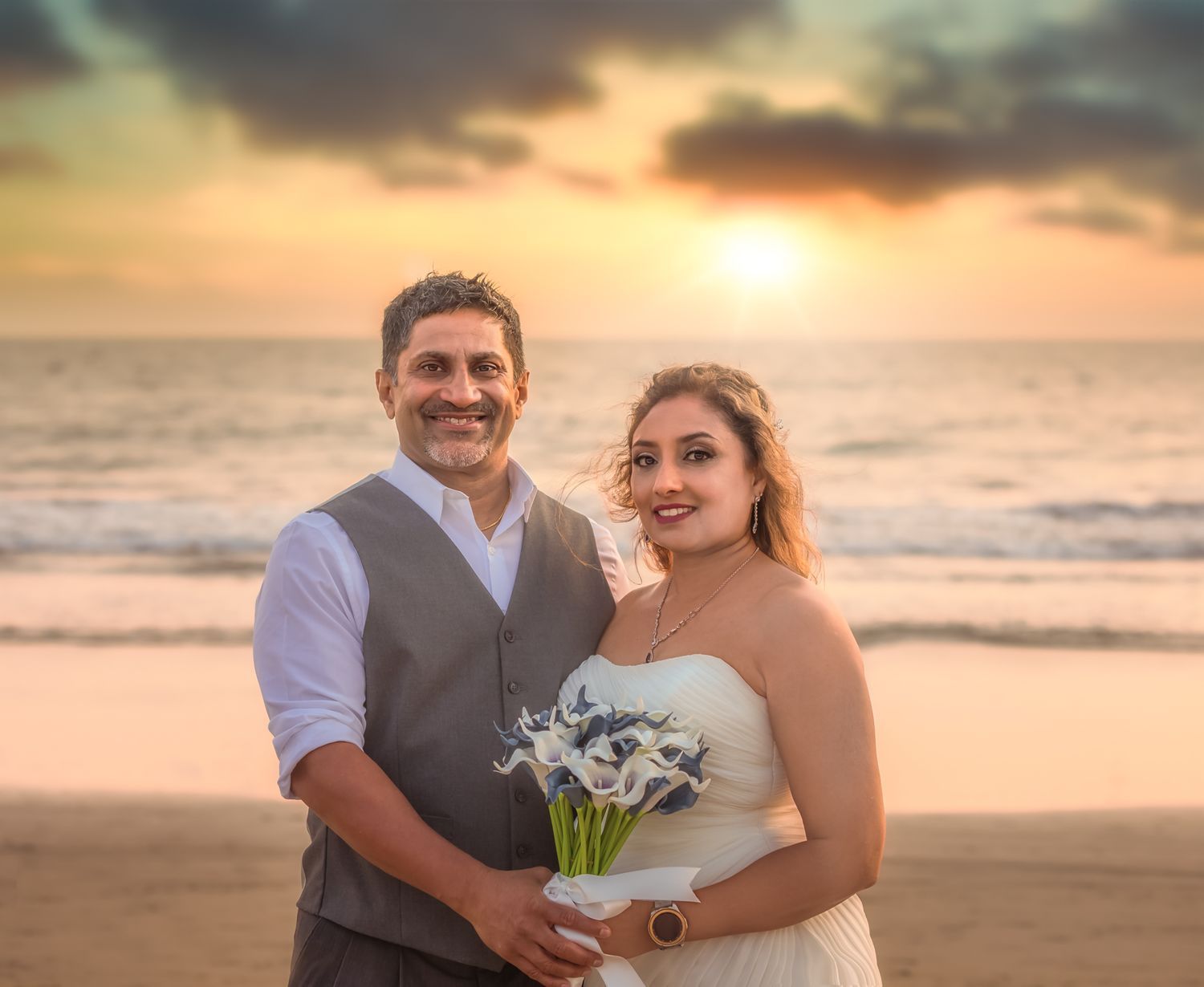 Newlywed couple standing on a beach at sunset, bride in white gown holding bouquet, groom in gray shirt.
