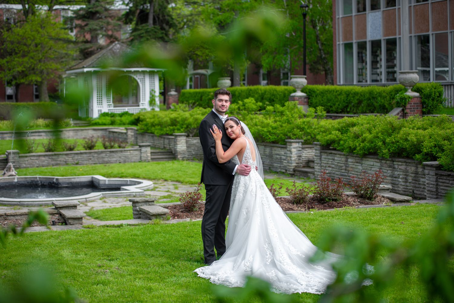 Bride and groom pose together in a lush garden with a fountain and gazebo on their wedding day.