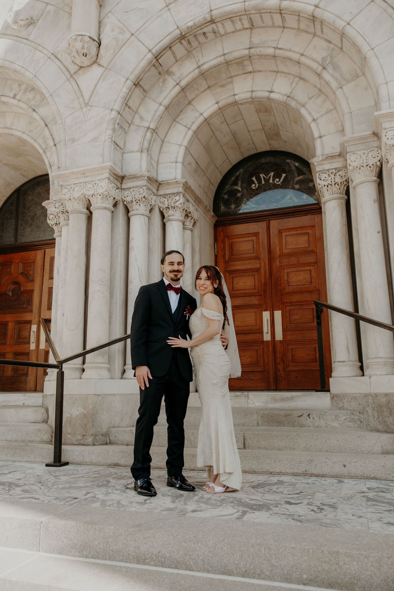 A bride and groom pose smiling in front of an ornate stone church entrance with large wooden doors.