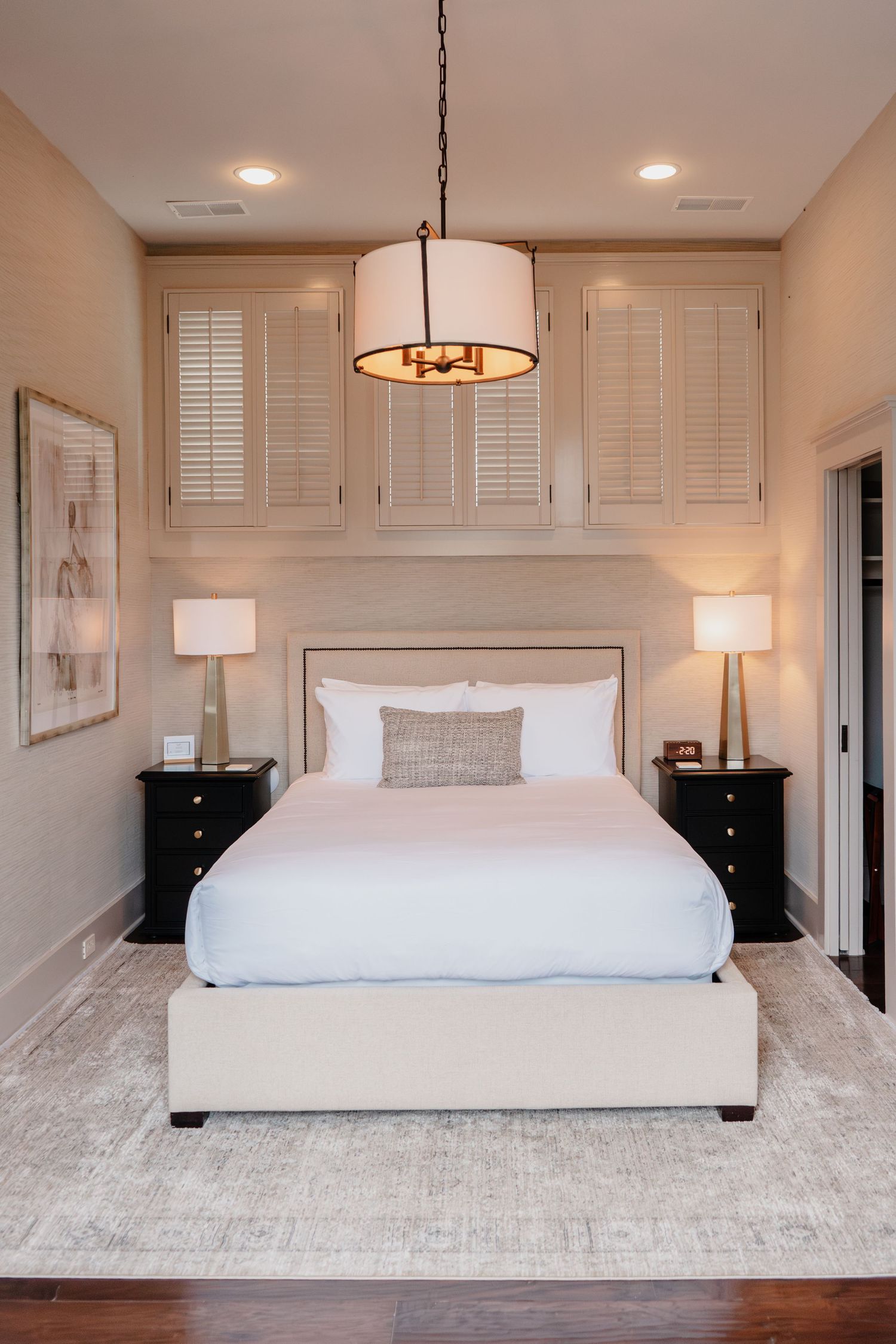 Elegant neutral bedroom with upholstered bed, black nightstands, pendant light, shuttered cabinets, and hardwood floors.