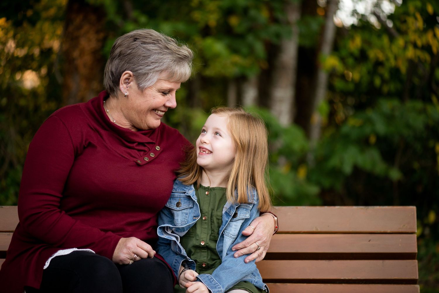 Grandmother and granddaughter sitting on a park bench, smiling at each other in an outdoor autumn setting.