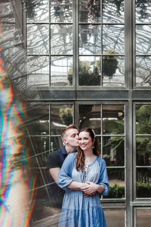 A couple embraces in front of a glass greenhouse conservatory with rainbow light prisms reflecting in the frame.