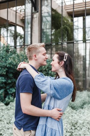 A couple in coordinating blue outfits embrace and share romantic moments in front of a greenhouse with lush greenery.