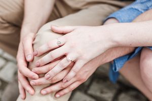 Close up of hands with engagement rings showing on fingers against a neutral background.