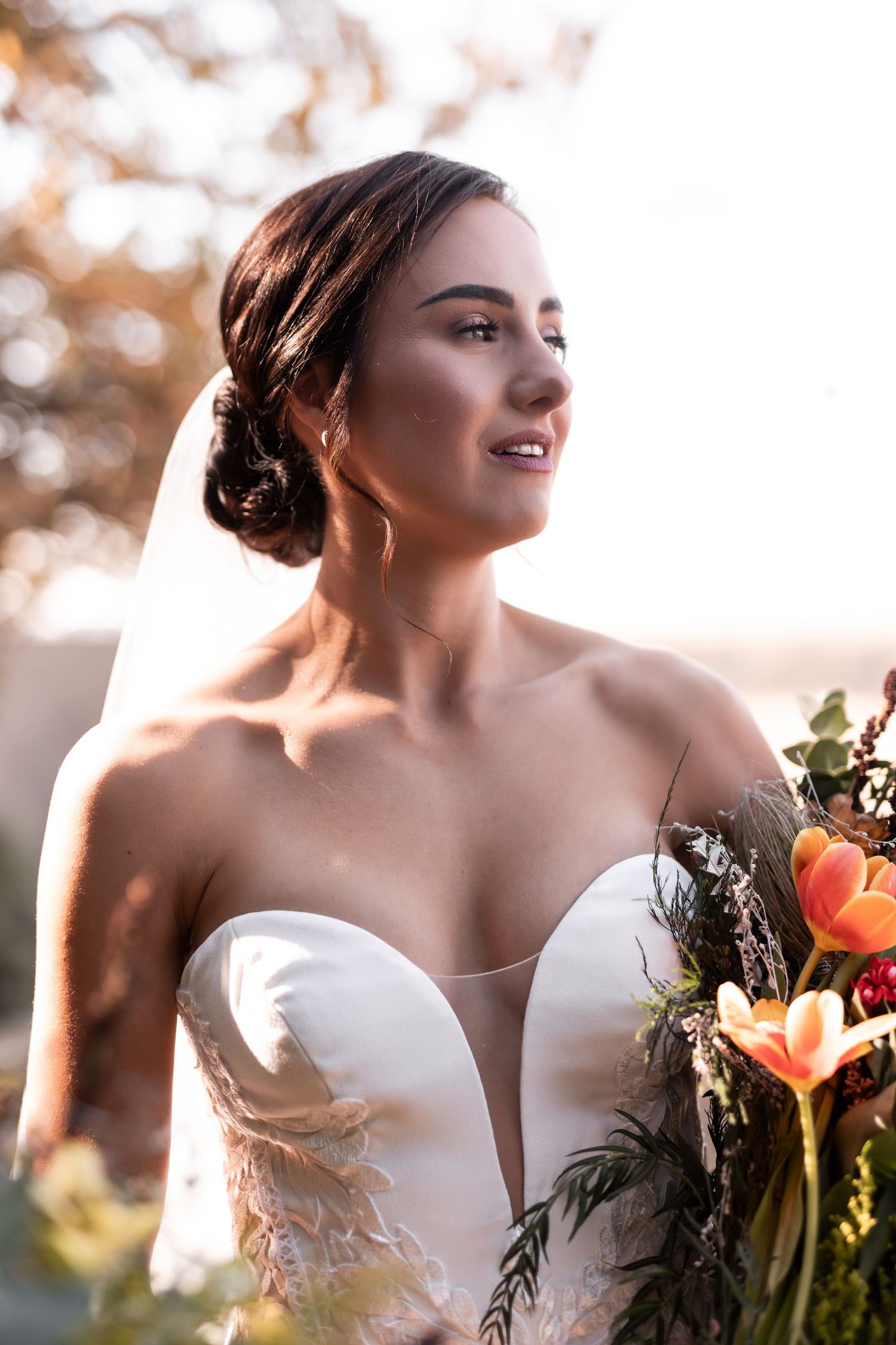 Elegant bride in a strapless gown holding a colorful floral bouquet in warm golden sunlight.