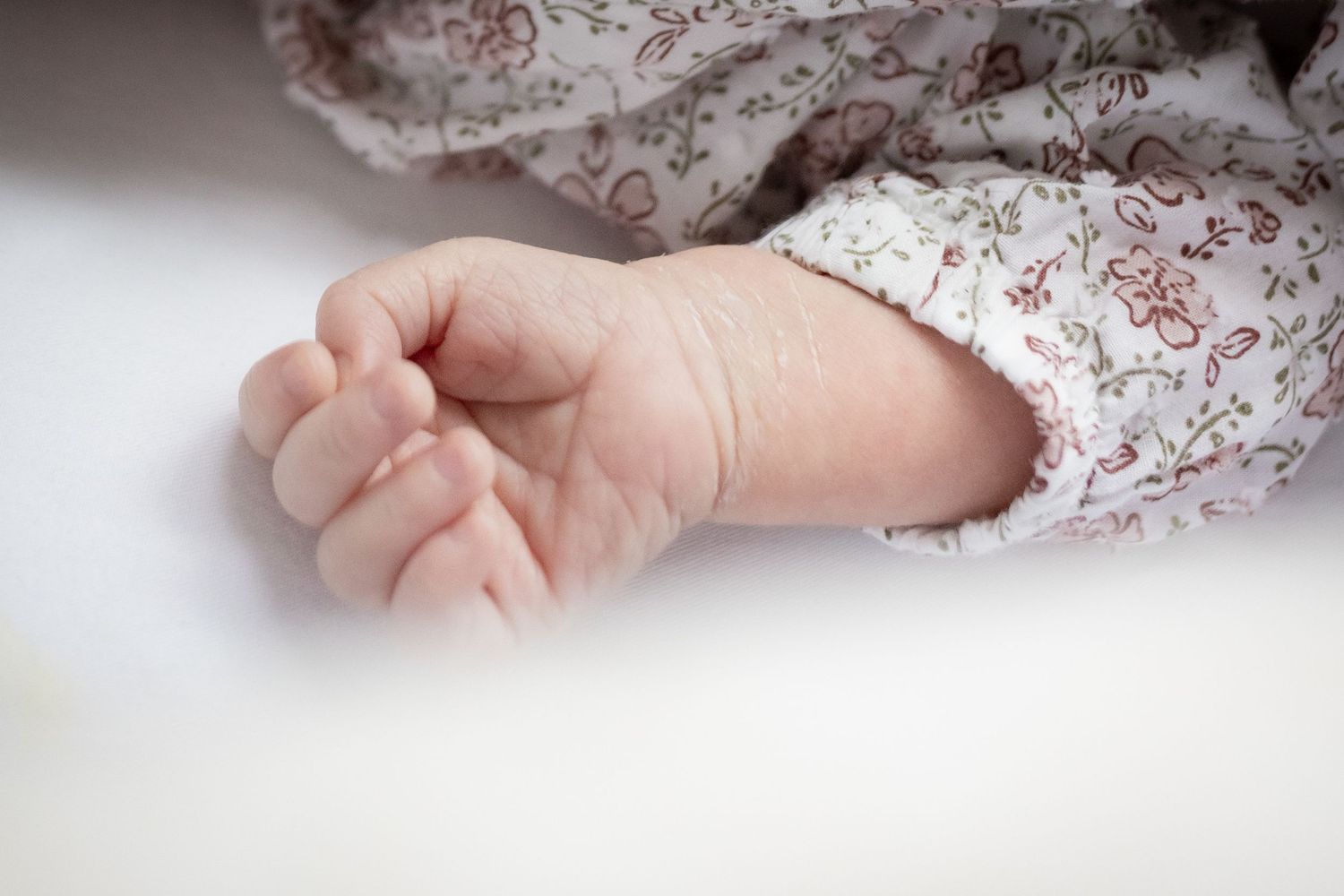 Close-up of newborn baby tiny clenched fist in floral outfit on white surface.