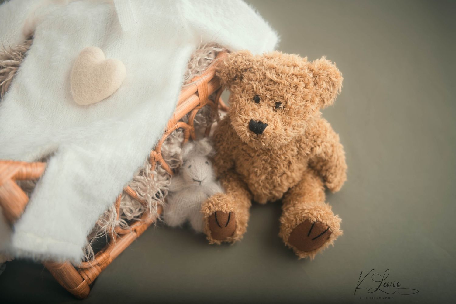 Fluffy brown teddy bear sitting beside a wicker basket with a white blanket and heart decoration. Newborn Photography by K Lewis Photography