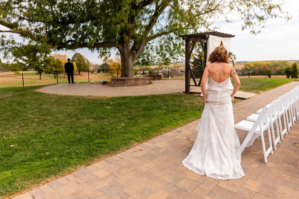 Bride in lace gown walks along brick path toward groom standing near a wooden arbor for their first look  at a Venue on the Ridge in Elkhorn