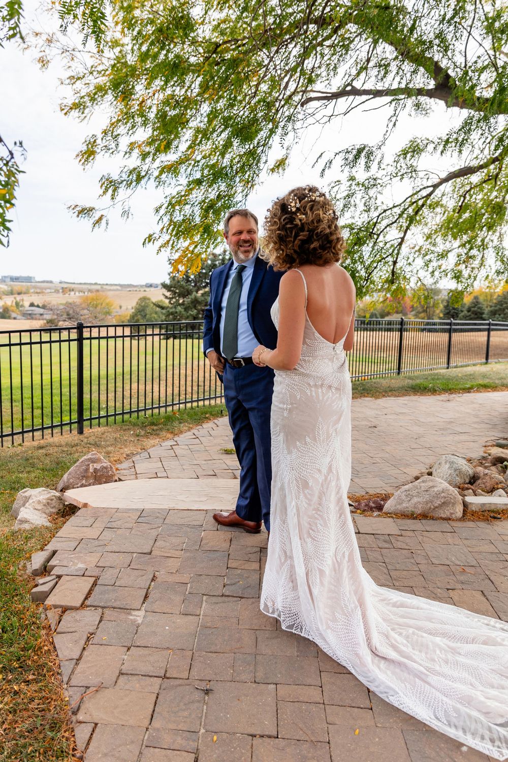 Groom in navy suit smiles at bride in backless lace wedding gown during an outdoor first look on a brick patio  at a Venue on the Ridge