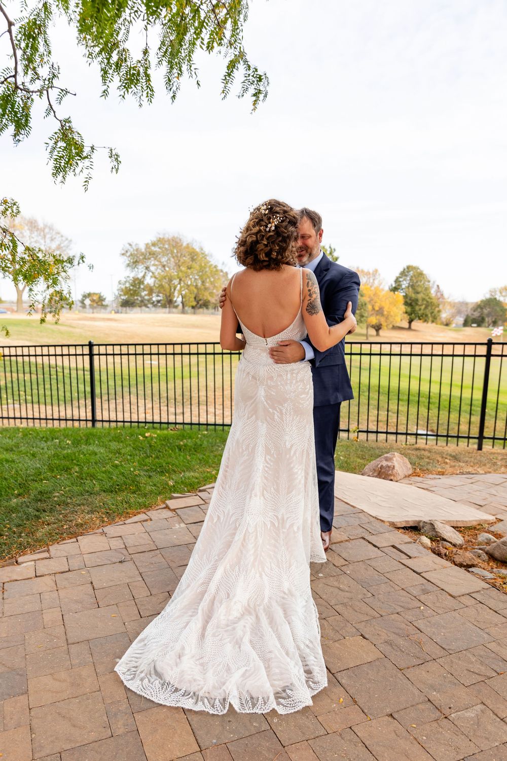 Bride in backless lace gown embraced by groom on a patio overlooking green landscape  at a Venue on the Ridge in Elkhorn, NE