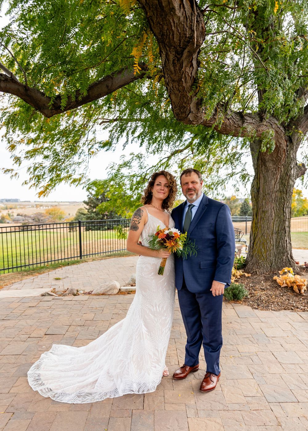 Bride and groom pose together under a large tree on a brick patio, bride holding a vibrant fall bouquet  at a Venue on the Ridge in Elkhorn,