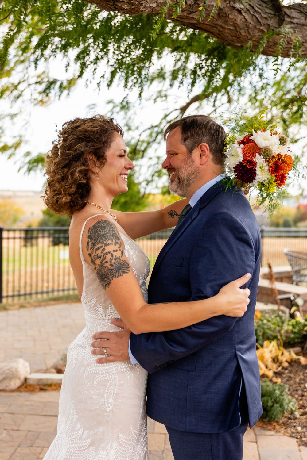 Smiling bride with tattoos and groom in navy suit embrace outdoors holding bouquet  at a Venue on the Ridge in Elkhorn, NE