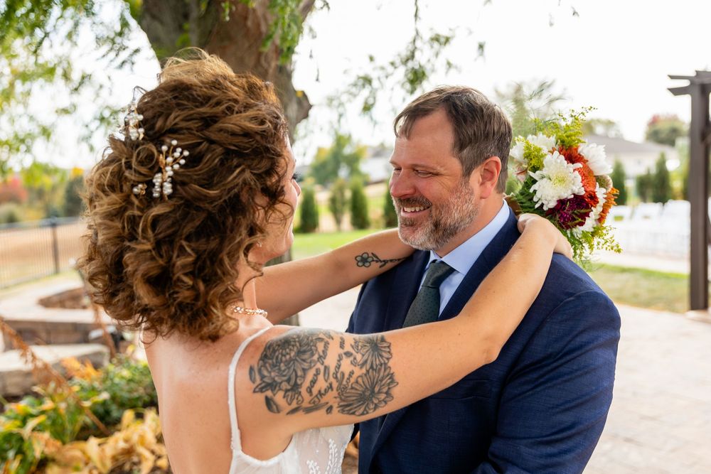 Bride with floral hair accessories and arm tattoo adjusts grooms navy suit collar during wedding first look  at a Venue on the Ridge